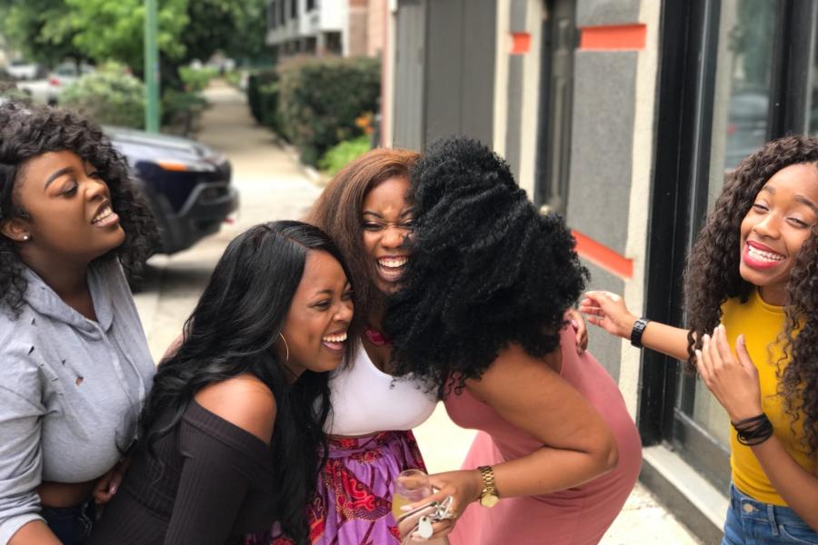 Five women laughing together while standing on a sidewalk