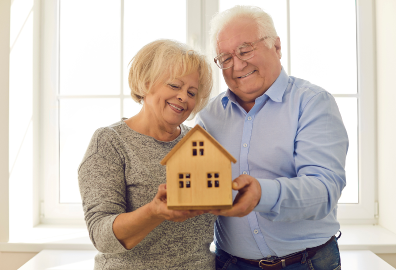 Elderly couple smiling in front of a window holding a small wooden house 