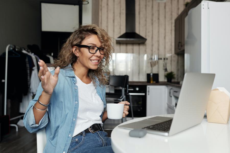 Woman sitting at kitchen table smiling at computer and gesturing while holding a coffee mug.