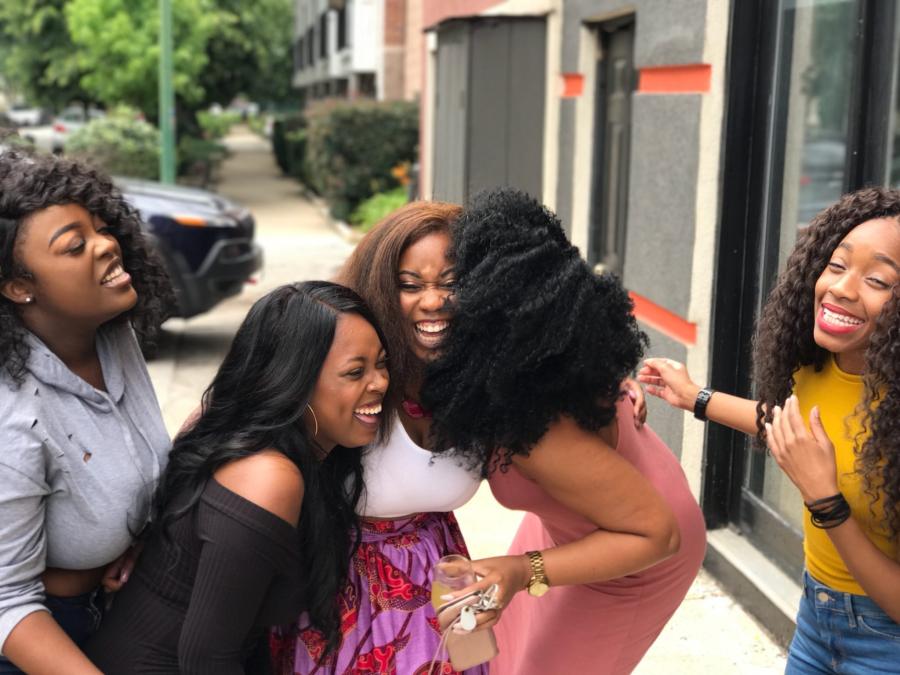 Five women laughing together while standing on a sidewalk