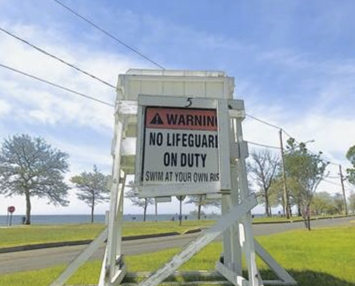 "No Lifeguard on Duty" sign. The white warning sign is outdoors on grass, near a waterfront